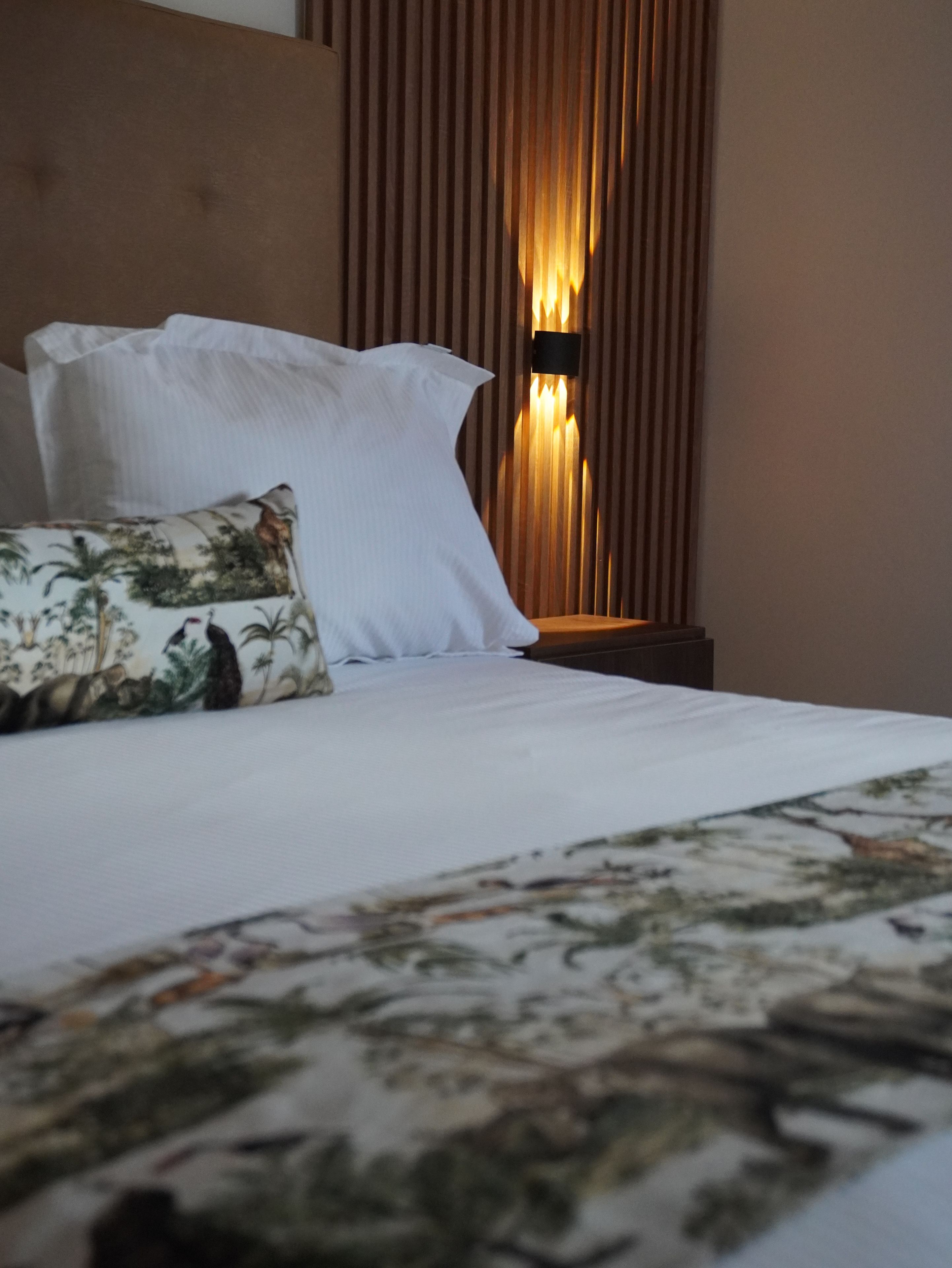 A bed with white bedding and a patterned throw, a pillow with a tropical print, and a wall-mounted light fixture.