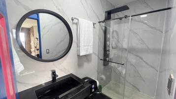 Un baño con pared de mármol, un espejo redondo, un lavabo negro, inodoro y ducha con mampara de vidrio.