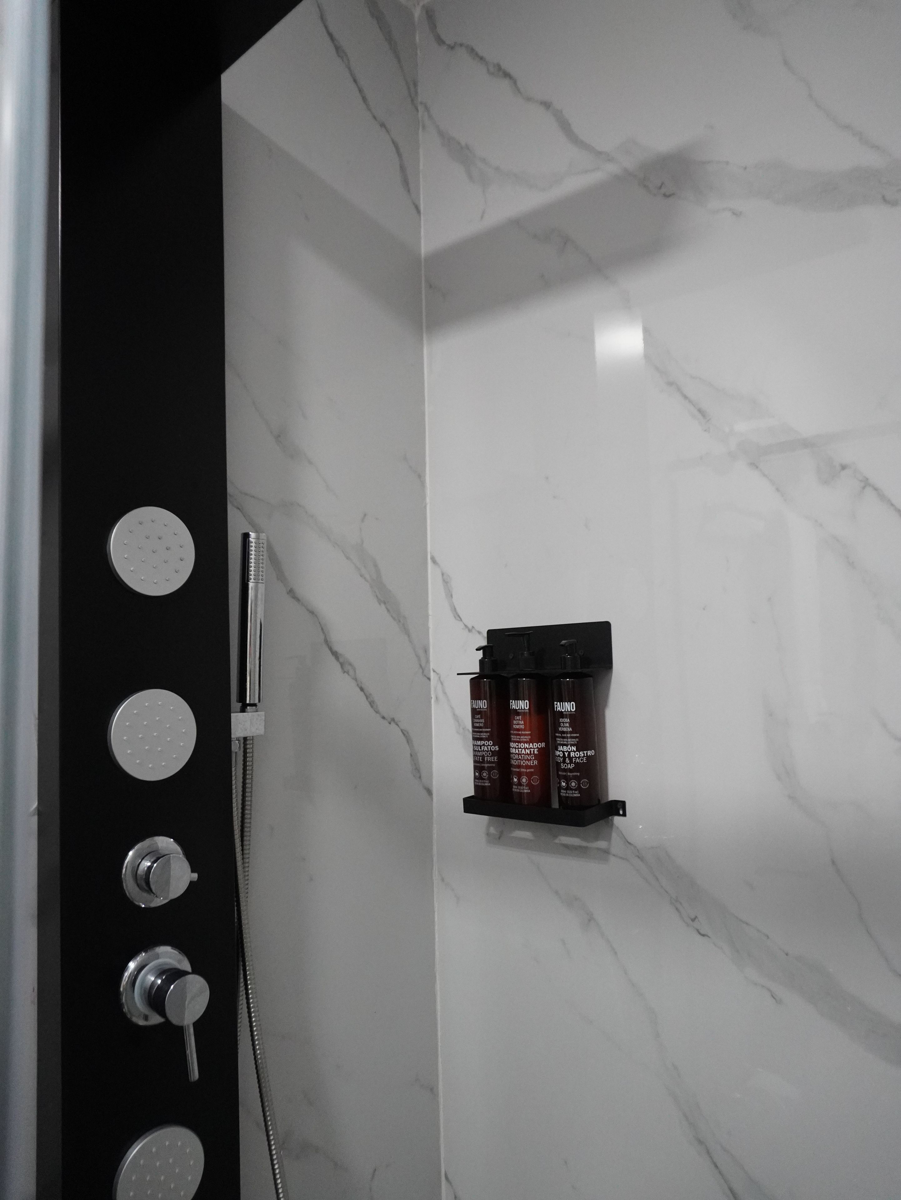 Une salle de bain avec un mur en marbre, un panneau noir doté de poignées rondes et une étagère contenant des flacons de produits de toilette.