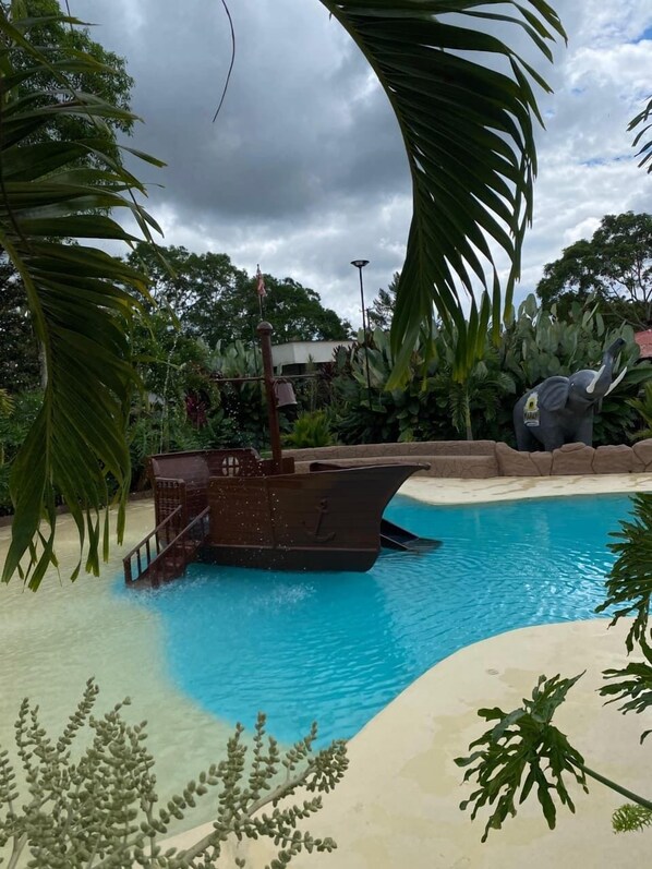 A swimming pool with a shipwreck sculpture, surrounded by tropical foliage and a cloudy sky. - MAHAWI RESORT (Calarca)