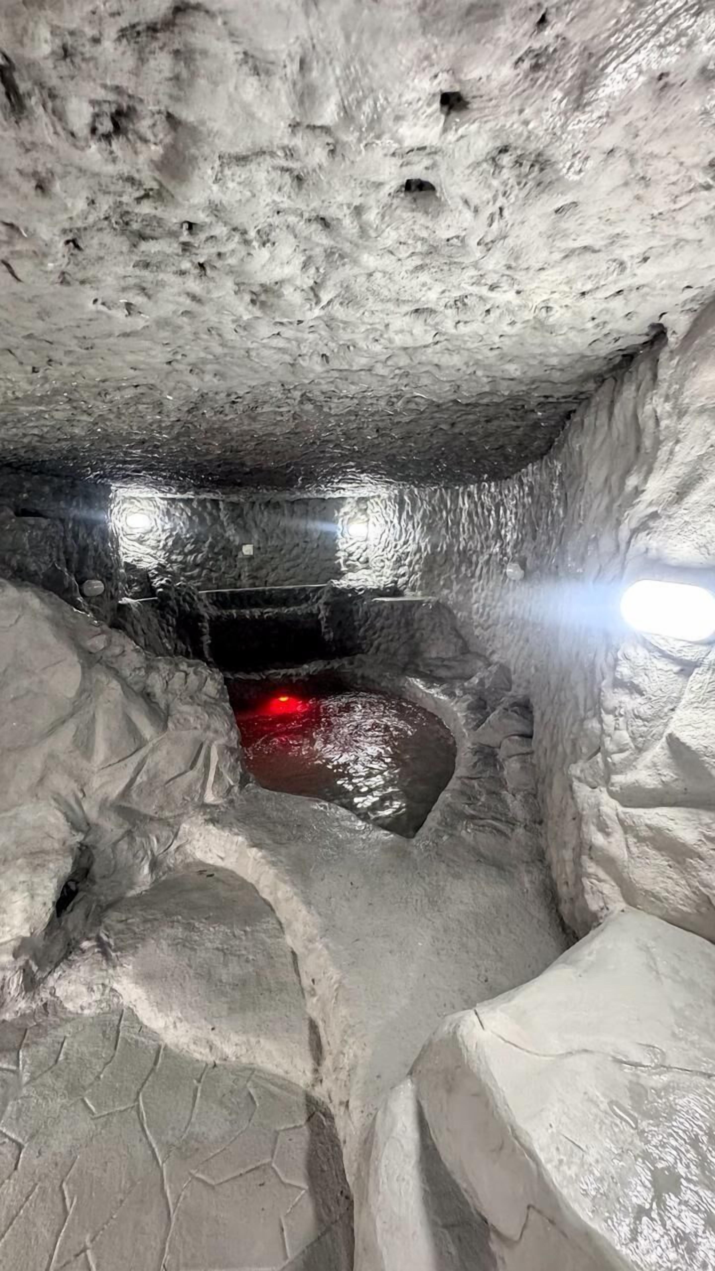 Un passage souterrain aux murs rugueux et texturés, illuminé d’une lumière rouge au fond.