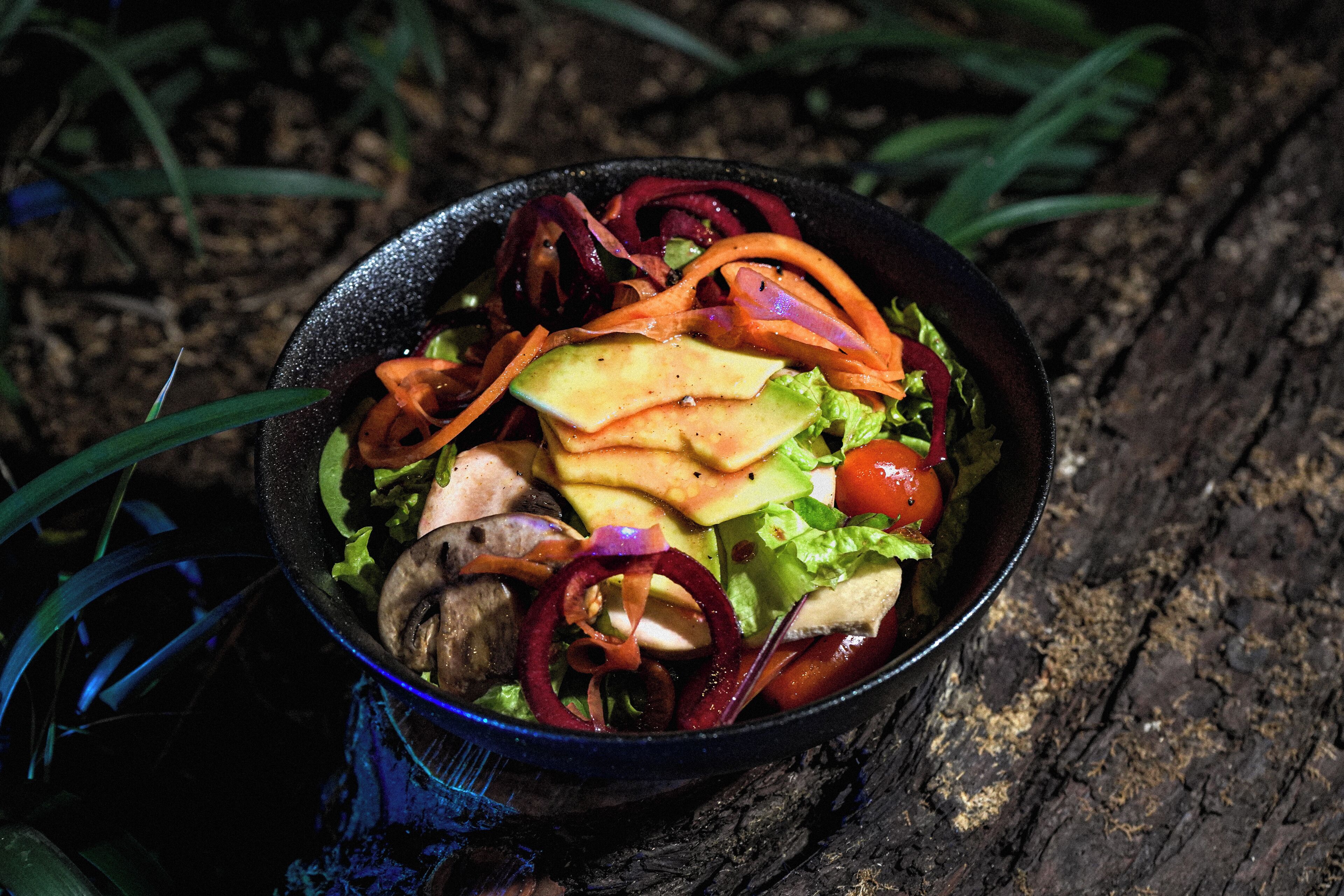 Un tazón de ensalada con diferentes verduras y una porción de queso sobre una superficie de madera.