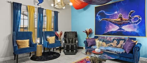 A living room with a blue sofa, a massage chair, a glass coffee table, and a large painting of a genie lamp.