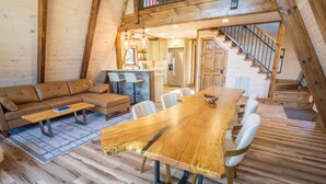 Een ruime loft met een hoge plafond, zichtbare houten balken, een grote houten eettafel, een zithoek en een trap naar een hoger niveau.