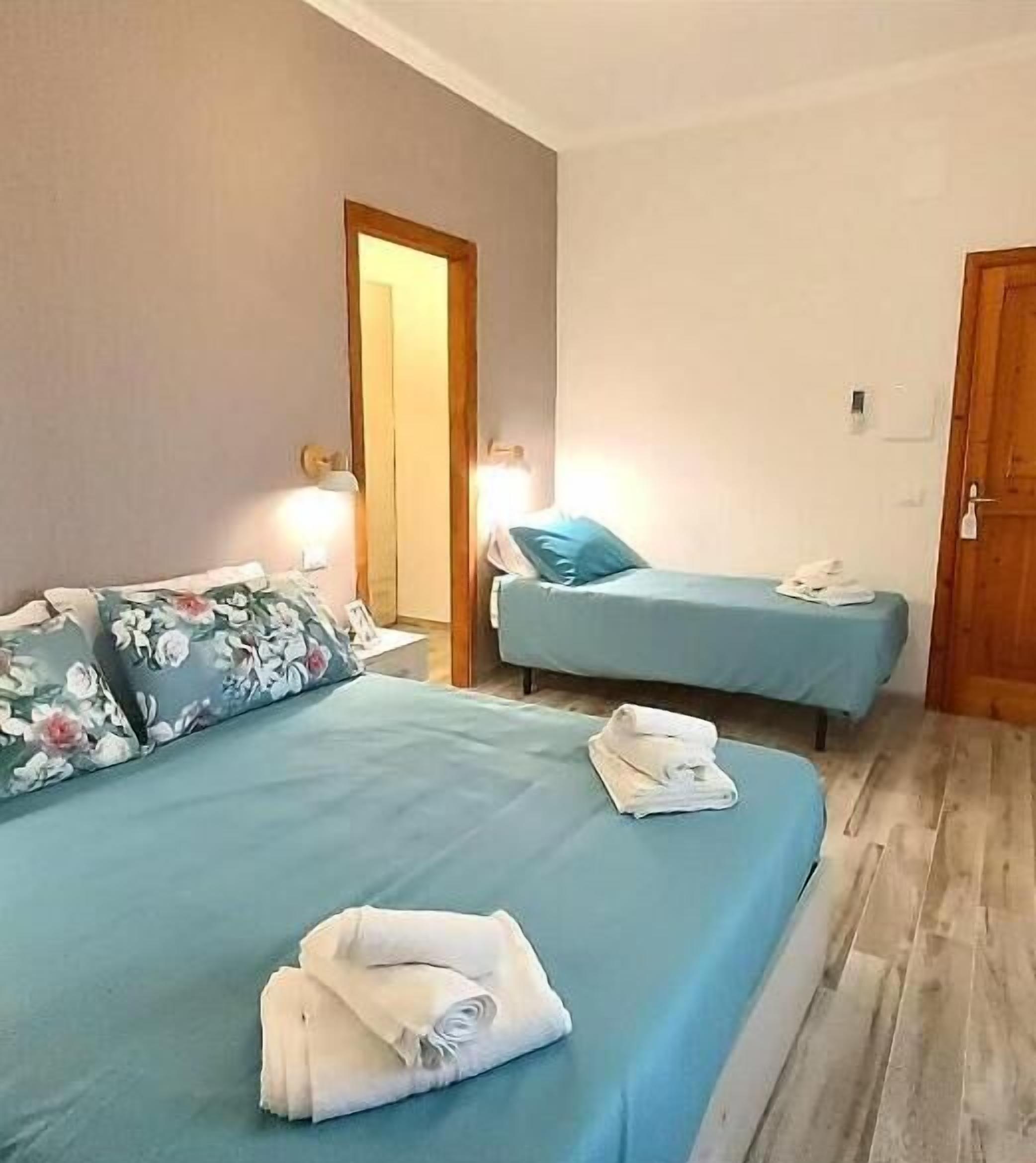 A hotel room with two beds, each with a blue bedspread and white pillows. There are folded towels on the beds, and a wooden door is visible in the background.