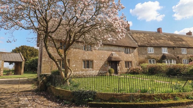 Une maison traditionnelle en pierre, avec un toit de chaume, une pelouse bien entretenue et une clôture en métal.