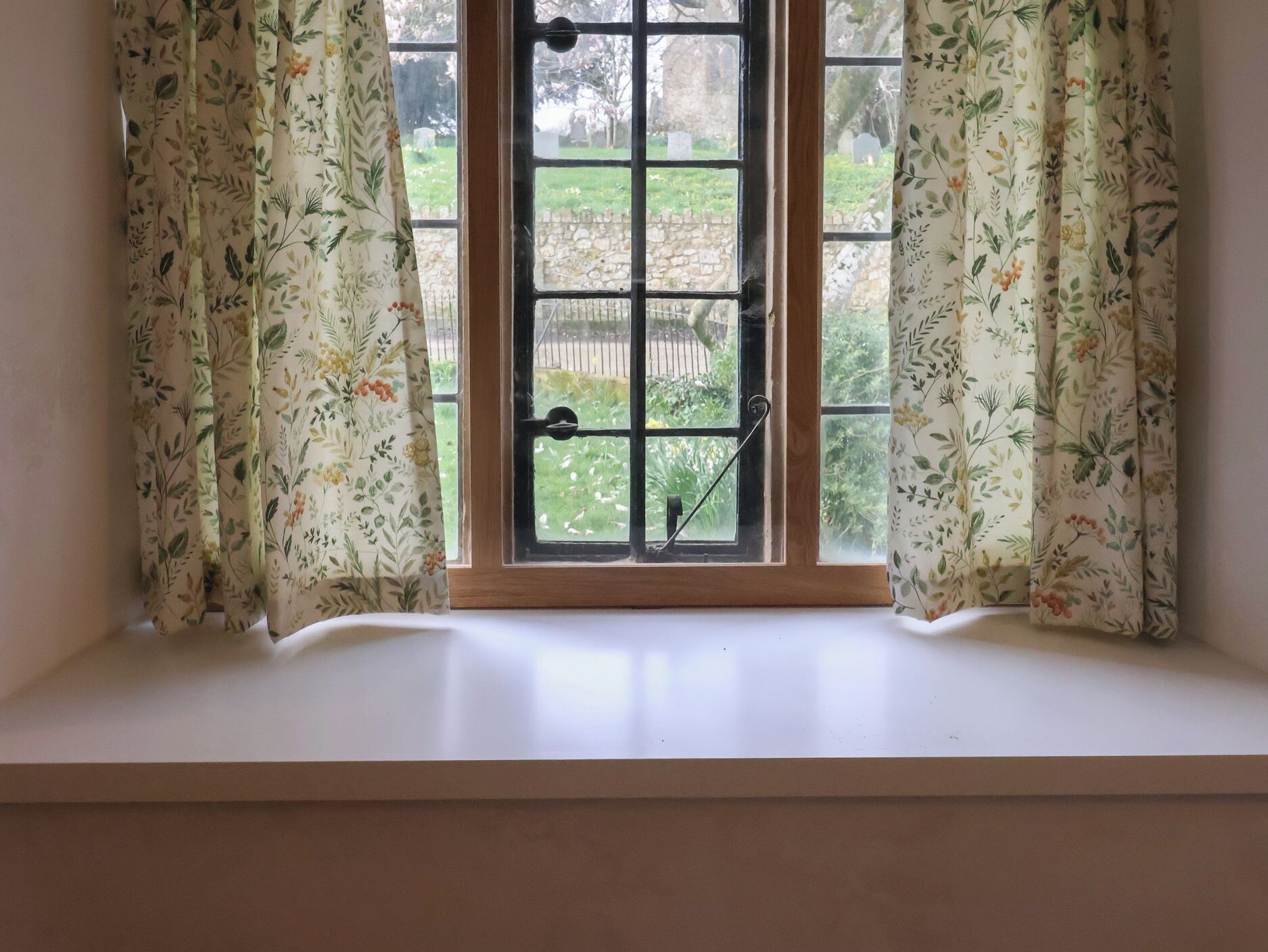 A window with floral curtains, a view of a cemetery, and a white window sill.