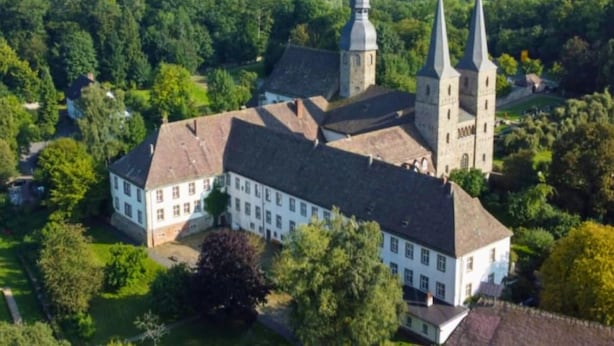 En stor, historisk byggnad med en central innergård, omgiven av frodig grönska och ett kuperat landskap.