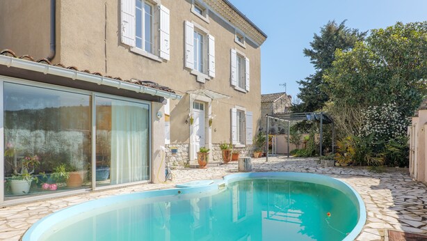 Ein Schwimmbecken mit Steinrand, ein Gebäude mit weißen Fensterläden und Kübelpflanzen.