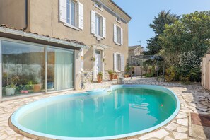 Una piscina con borde de piedra, un edificio con contraventanas blancas y plantas en macetas.