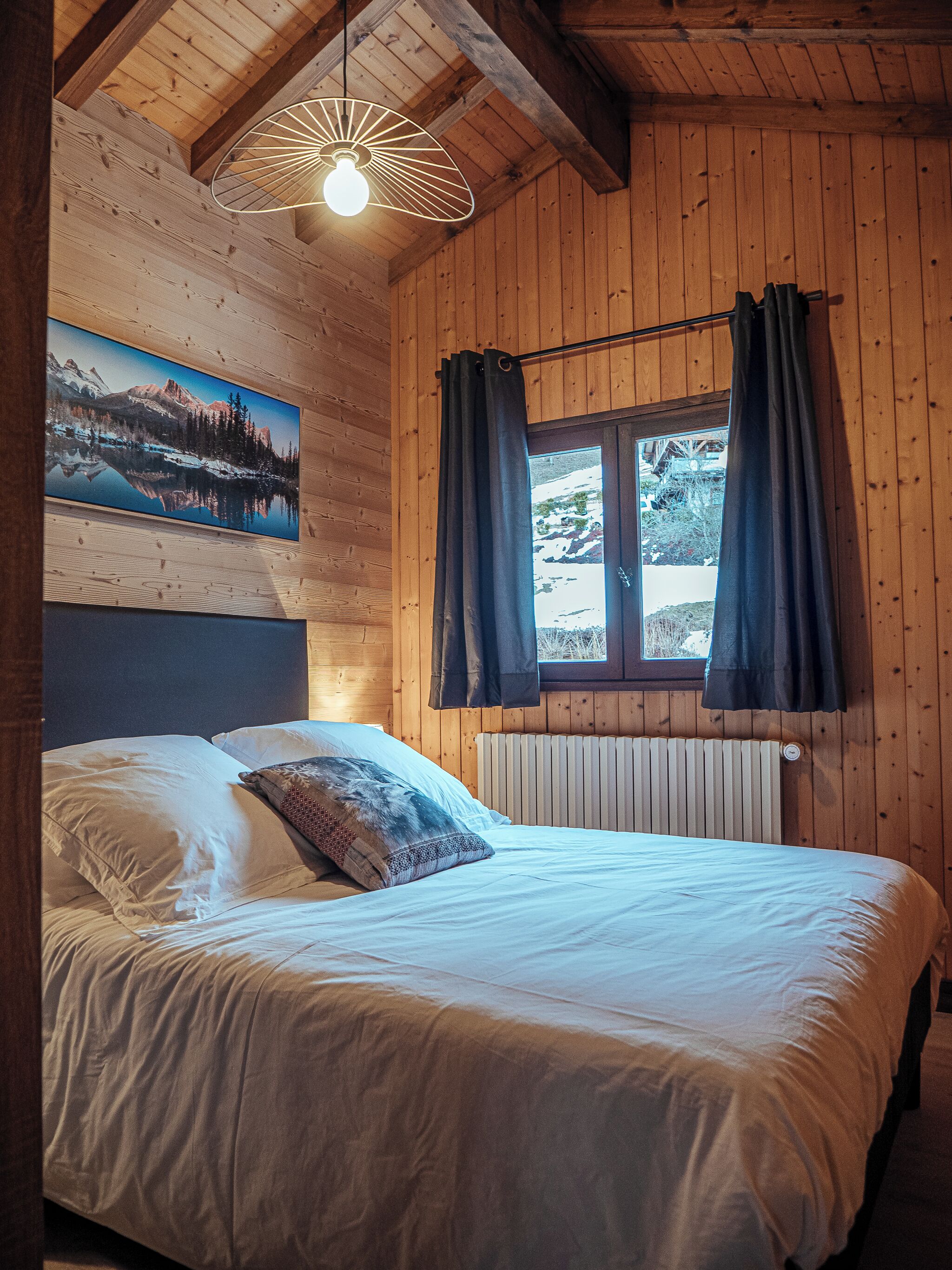 Habitació amb panells de fusta, un llit, una finestra amb cortines, un radiador i un quadre penjat a la paret.