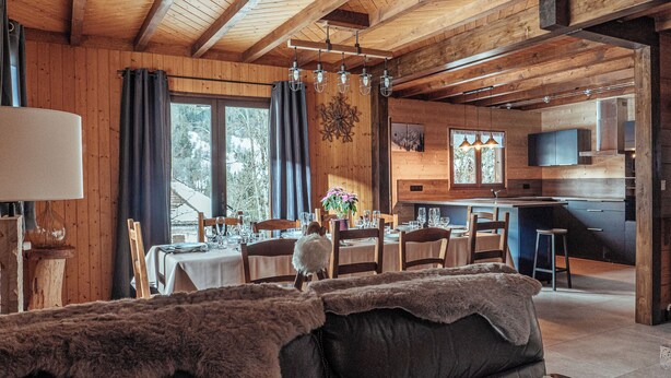Interior acolhedor de uma cabana de madeira, com área de jantar, cozinha e janelas amplas com vista para a paisagem nevada.