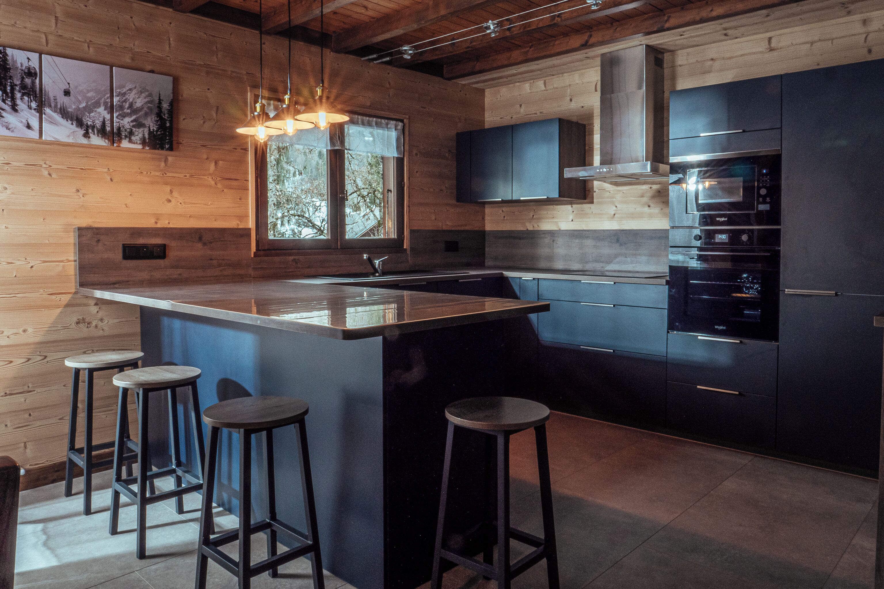 Una cocina moderna con una isla grande, taburetes de bar, horno empotrado y una ventana con vistas a árboles.