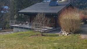 Uma cabana de madeira com telhado inclinado, um pequeno alpendre e vista para montanhas nevadas.
