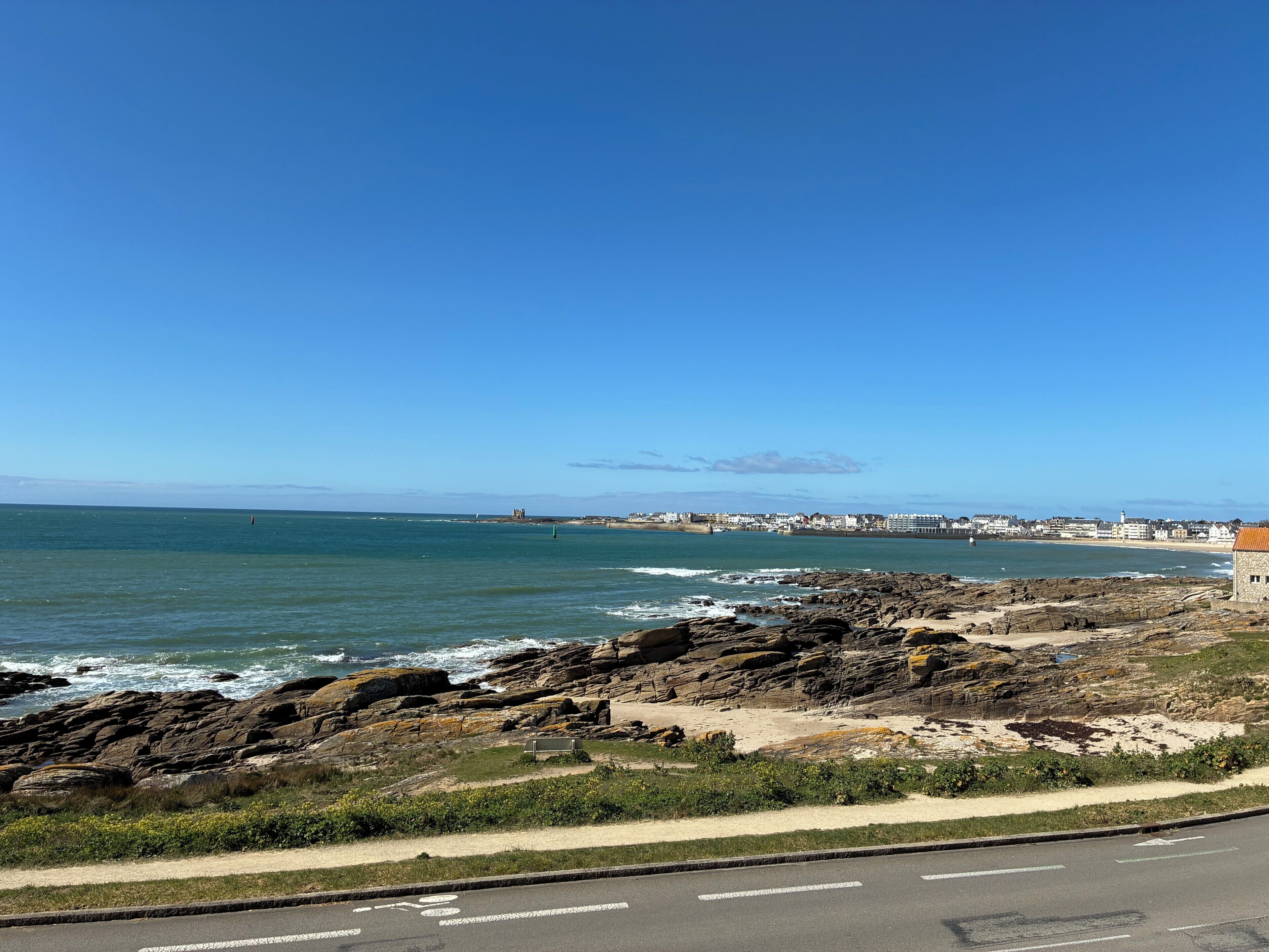 Une route côtière bordée d’une côte rocheuse, sous un ciel bleu dégagé, avec des bâtiments visibles au loin le long de la côte.