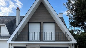 Et moderne hus i to etager med skrå tag, to garageporte og en balkon med gelænder.