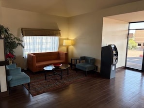 En hotellobby med en soffa, fåtöljer, ett litet bord, en lampa, en automater och ett stort fönster.