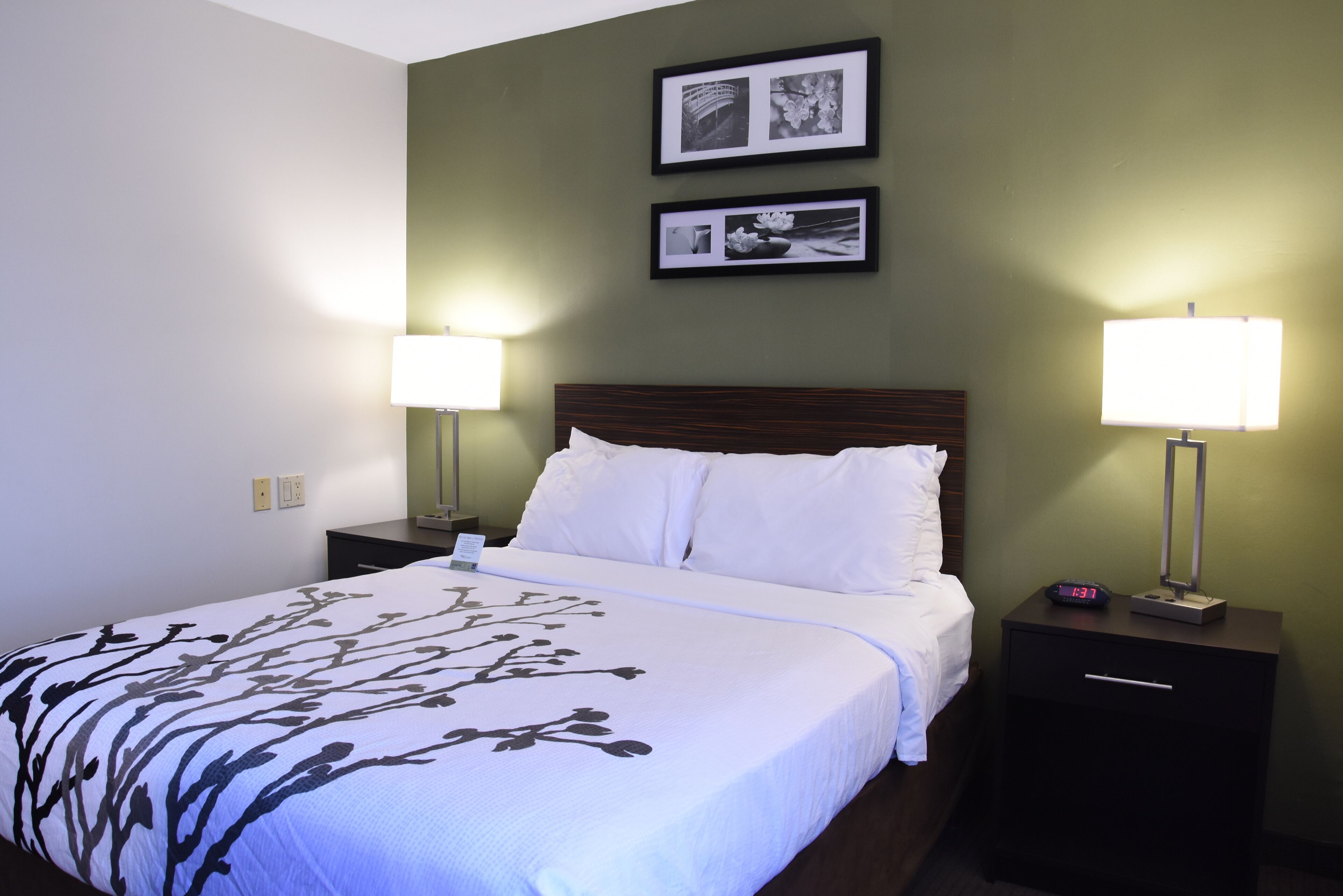 A neatly made bed with white linens and a floral-patterned duvet, flanked by two bedside tables with lamps, each table featuring a digital clock.