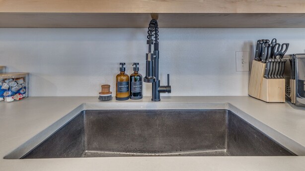 Una cocina con un lavamanos de acero inoxidable, estantes de madera arriba y diversos utensilios de cocina y botellas en la encimera.