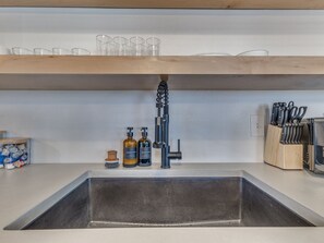 Eine Küche mit Spüle aus Edelstahl, Holzregalen darüber und verschiedenen Küchenutensilien und Flaschen auf der Arbeitsfläche.