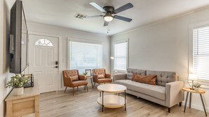 Una sala de estar con un ventilador de techo, un sofá, dos sillones, una mesa de centro y un televisor montado en la pared.