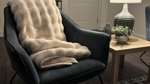 Een zwarte fauteuil met een witte plaid, een houten bijzettafeltje met een lamp en een kleine plant, een kamerbreed tapijt en een deur op de achtergrond.