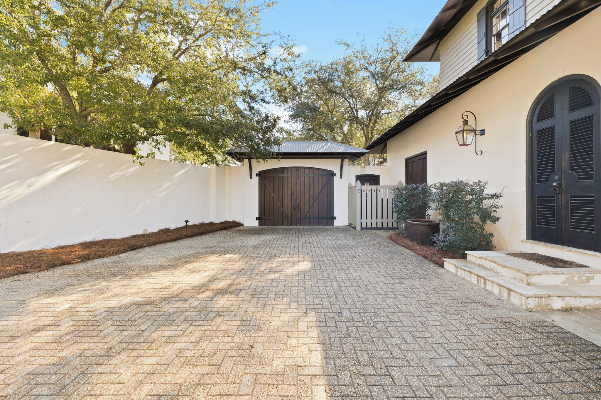 A paved driveway leading to a gated entrance with a large wooden door, a white picket fence, and a potted plant.