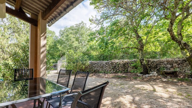 Un área de comedor al aire libre techada, con una mesa y sillas, rodeada de árboles y un muro de piedra.