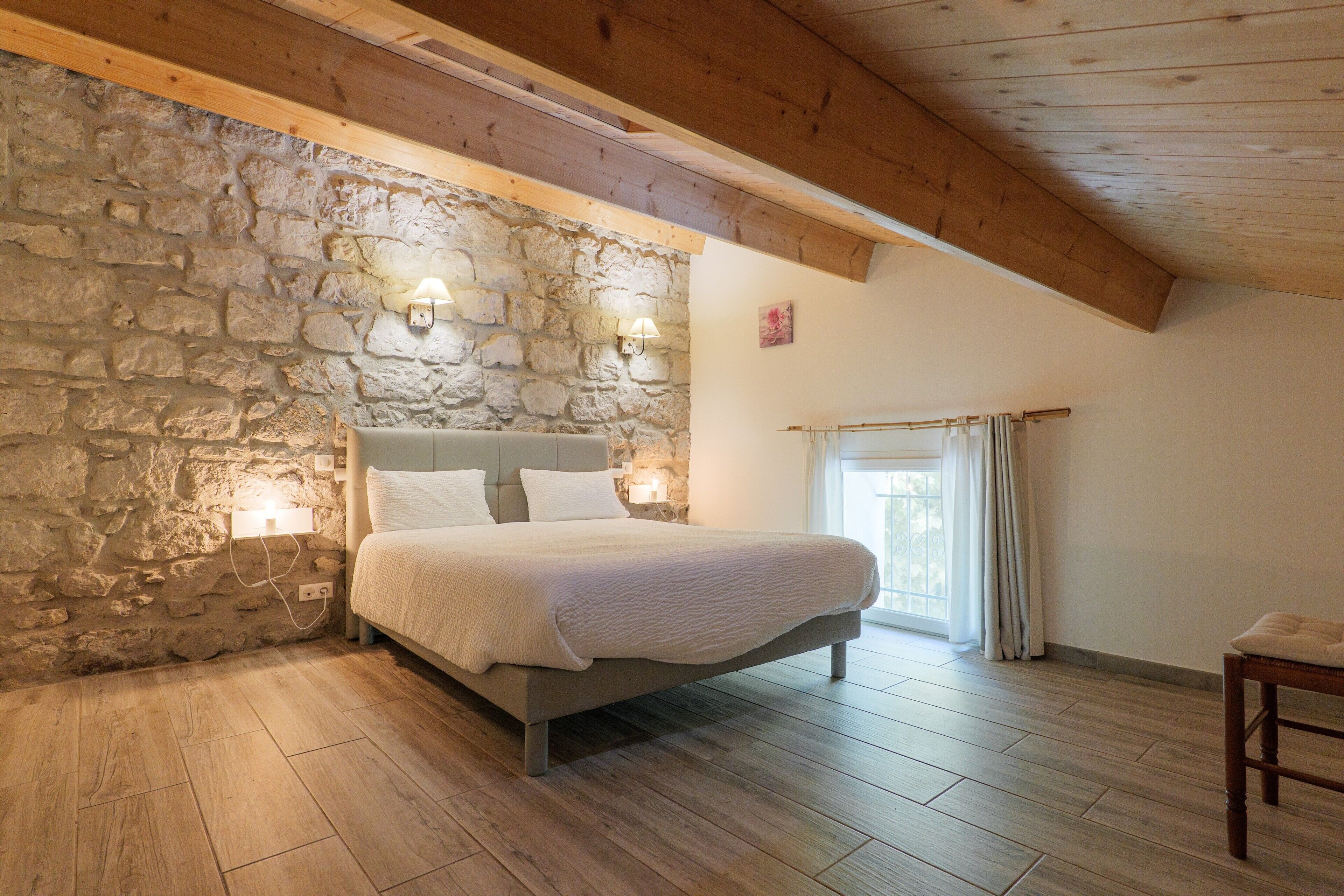 Un dormitorio con pared de piedra, suelo de madera, una cama con ropa de cama blanca y dos luces fijadas a la pared.