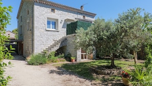 Une maison en pierre avec un escalier menant à un deuxième étage, entourée de verdure et bordée d’un chemin de gravier.