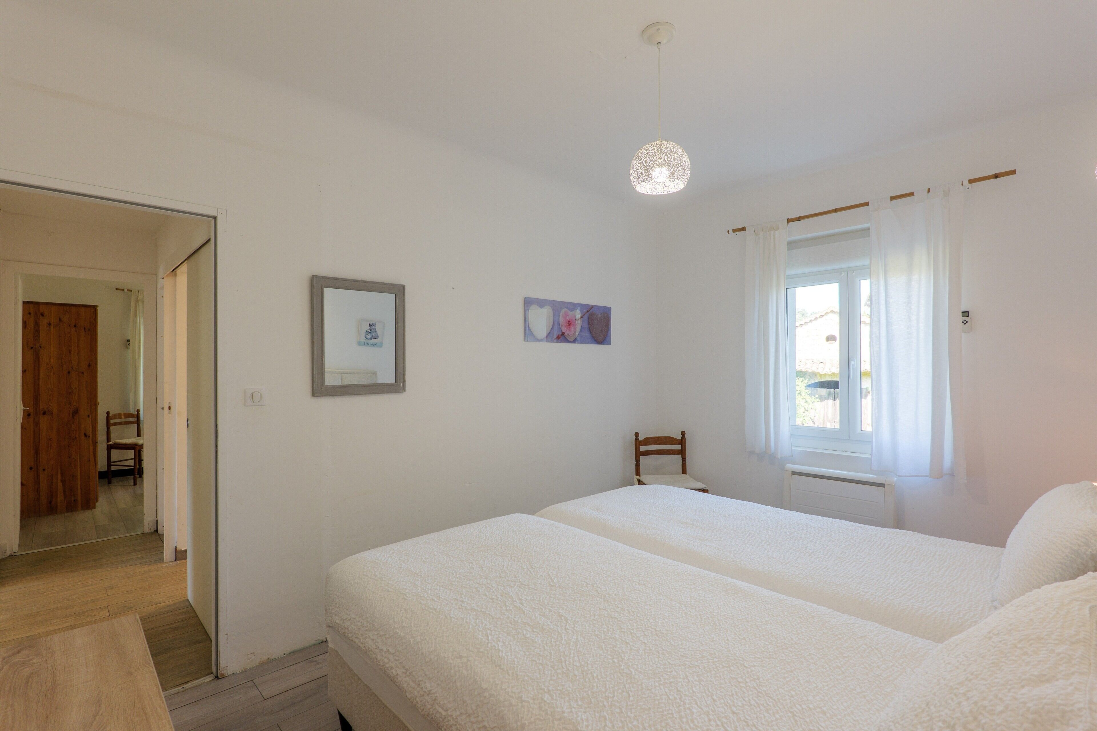 Quarto com duas camas, porta de madeira, quadro emoldurado e janela com cortinas.
