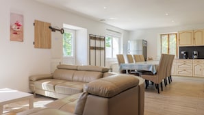 Un salon avec un canapé beige, une table à manger avec des chaises, un coin cuisine avec des placards et une fenêtre donnant sur un espace verdoyant.