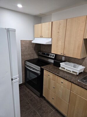 A compact kitchen with a built-in oven, a stovetop, and a microwave. There is a white refrigerator on the left, and a sink with a countertop on the right. The kitchen has wooden cabinets and a tiled backsplash.