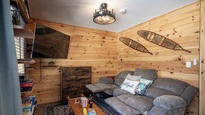 L’intérieur chaleureux d’un chalet avec un décor en bois, un canapé agrémenté d’un coussin « pour les invités », un téléviseur à écran plat et un ventilateur de plafond.