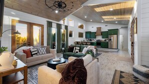 Amplia sala de estar con sofá, mesa de centro y chimenea. Hay una cocina con gabinetes verdes y un refrigerador. El espacio tiene techos de madera y ventanas grandes.