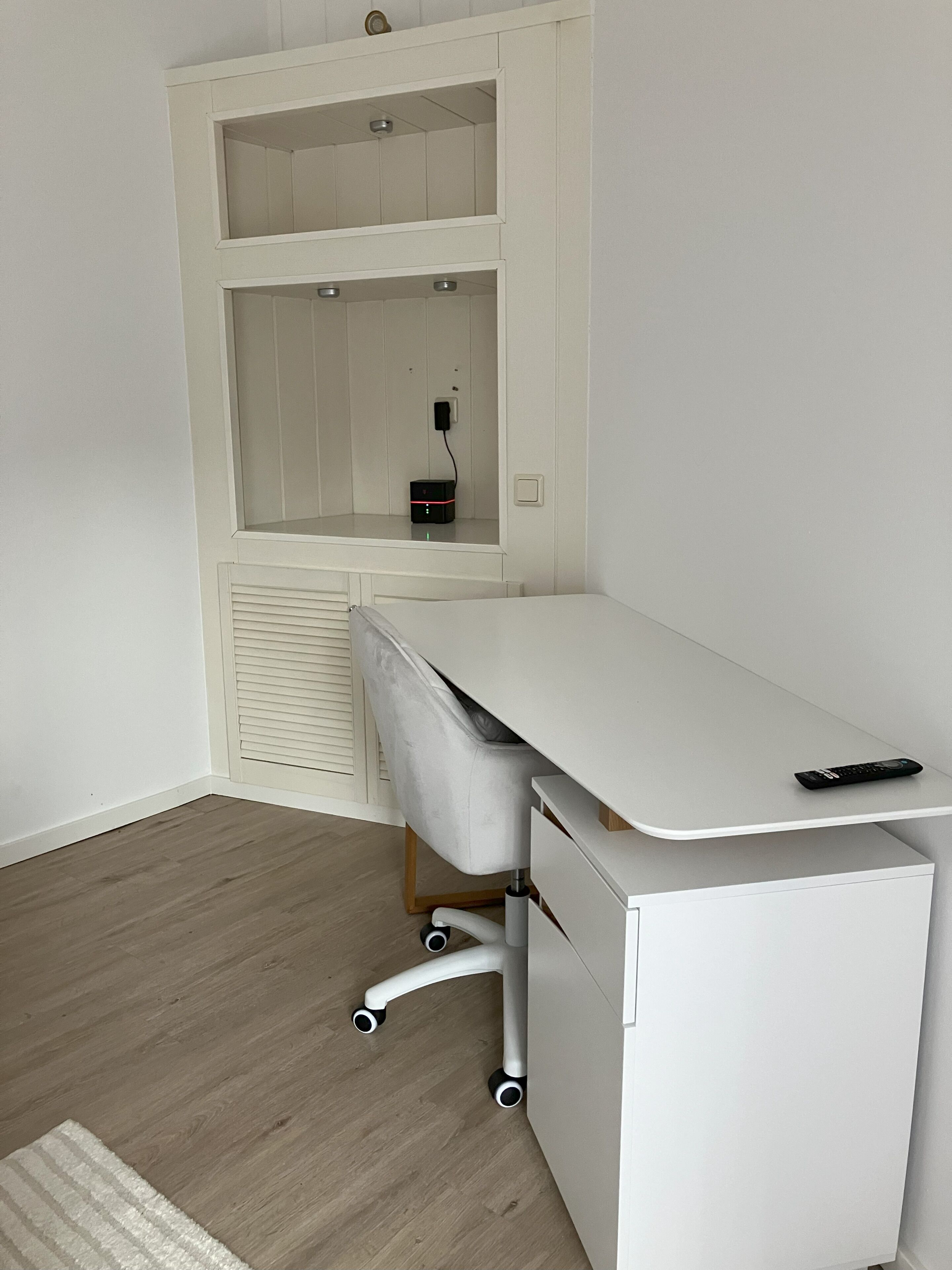 A white desk with a remote control on top, positioned next to a white cabinet with a shelf above.
