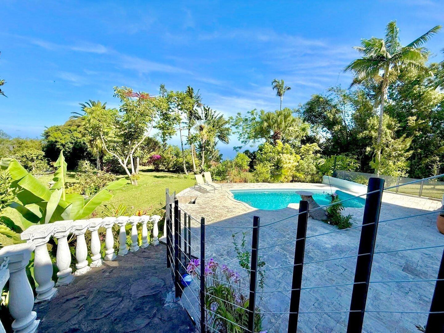 A pool area with a stone deck, surrounded by tropical plants and palm trees, overlooking a body of water.