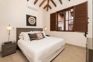 Un dormitorio con cama, mesitas de noche, aire acondicionado de pared, ventana con persianas de madera y un elemento decorativo en la pared.