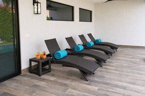 Een rij comfortabele stoelen met blauwe kussens, een klein tafeltje met drankjes, en een uitzicht op een zwembad door een glazen deur.