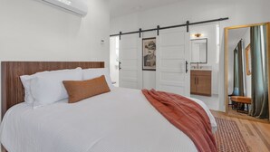 Une chambre avec un grand lit, une tête de lit en bois, un climatiseur blanc, une porte coulissante, une salle de bain avec un lavabo et un miroir, et un grand miroir avec un rideau vert.