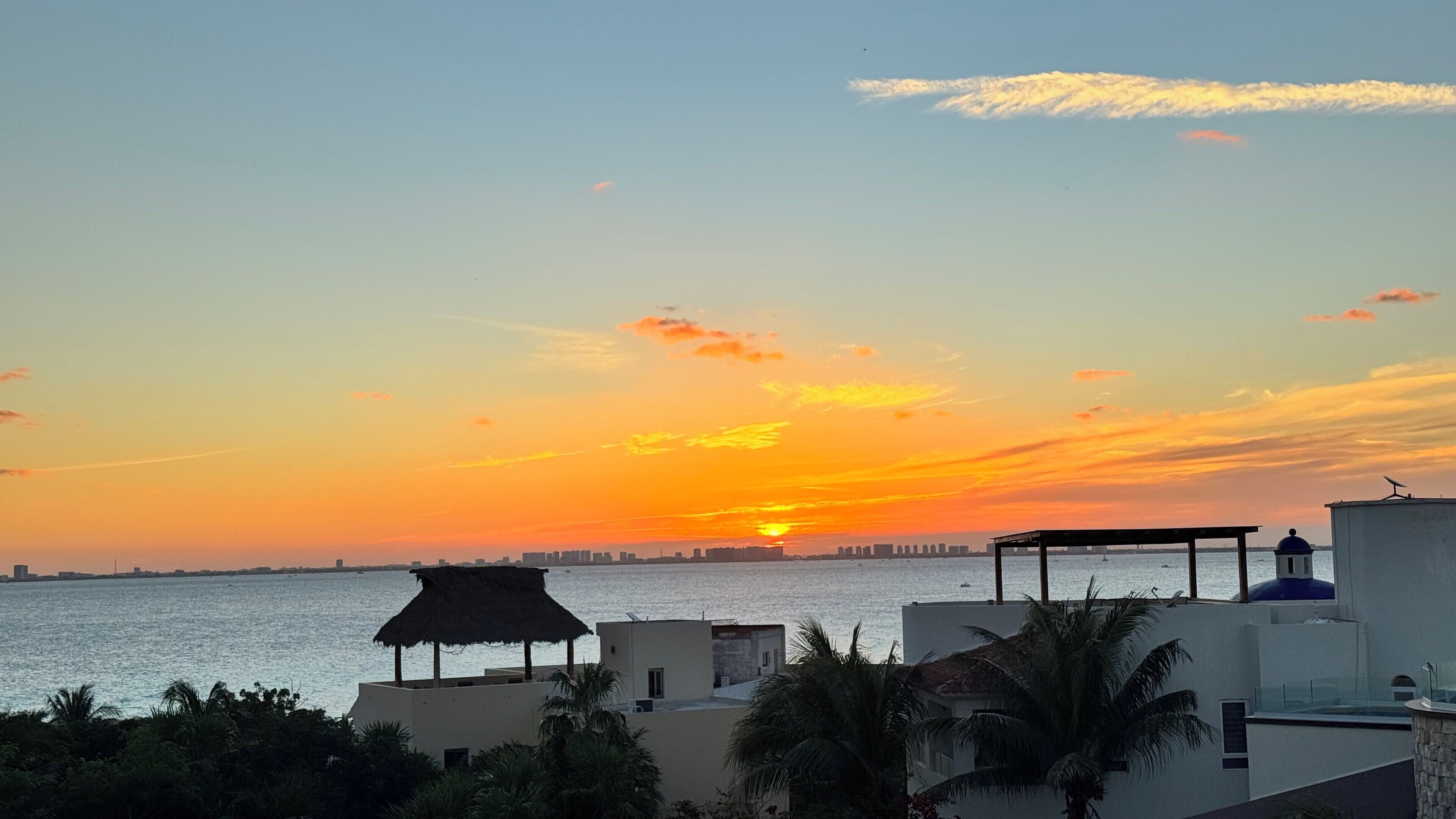 A sunset view over a body of water with buildings and a thatched-roof structure in the foreground.