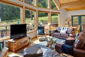 A spacious living room with a large TV, comfortable seating, and large windows offering a view of the outdoors.