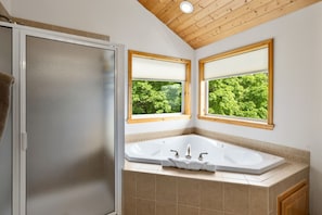 A bathroom with a large bathtub, two windows with blinds, and a shower door.