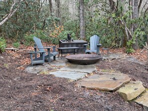 A rustic outdoor seating area with Adirondack chairs, a stone fire pit, and a wooden table. - Creekside Mountain Retreat 2+Acres Near Cashiers/Highlands (Cullowhee)