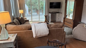Ein gemütliches Wohnzimmer mit einer Couch, einem Holzstuhl, einem Tisch, einem Fernseher und einem großen Fenster mit Vorhängen.