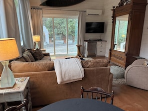 A cozy living room with a sofa, a wooden chair, a table, a TV, and a large window with curtains. - Creekside Mountain Retreat 2+Acres Near Cashiers/Highlands (Cullowhee)