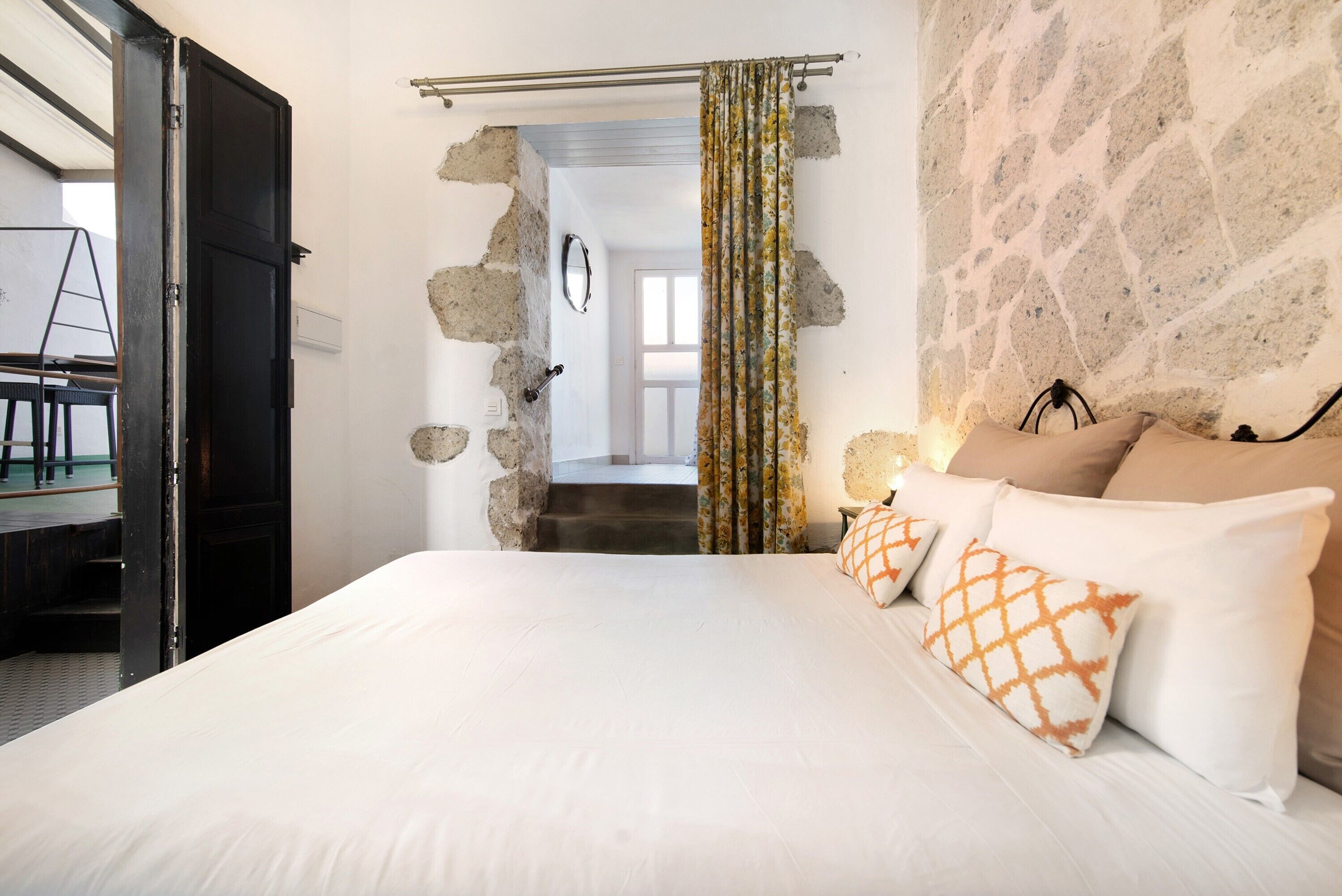 A bedroom with a bed, pillows, a curtain, a stone wall, and a doorway leading to another room.