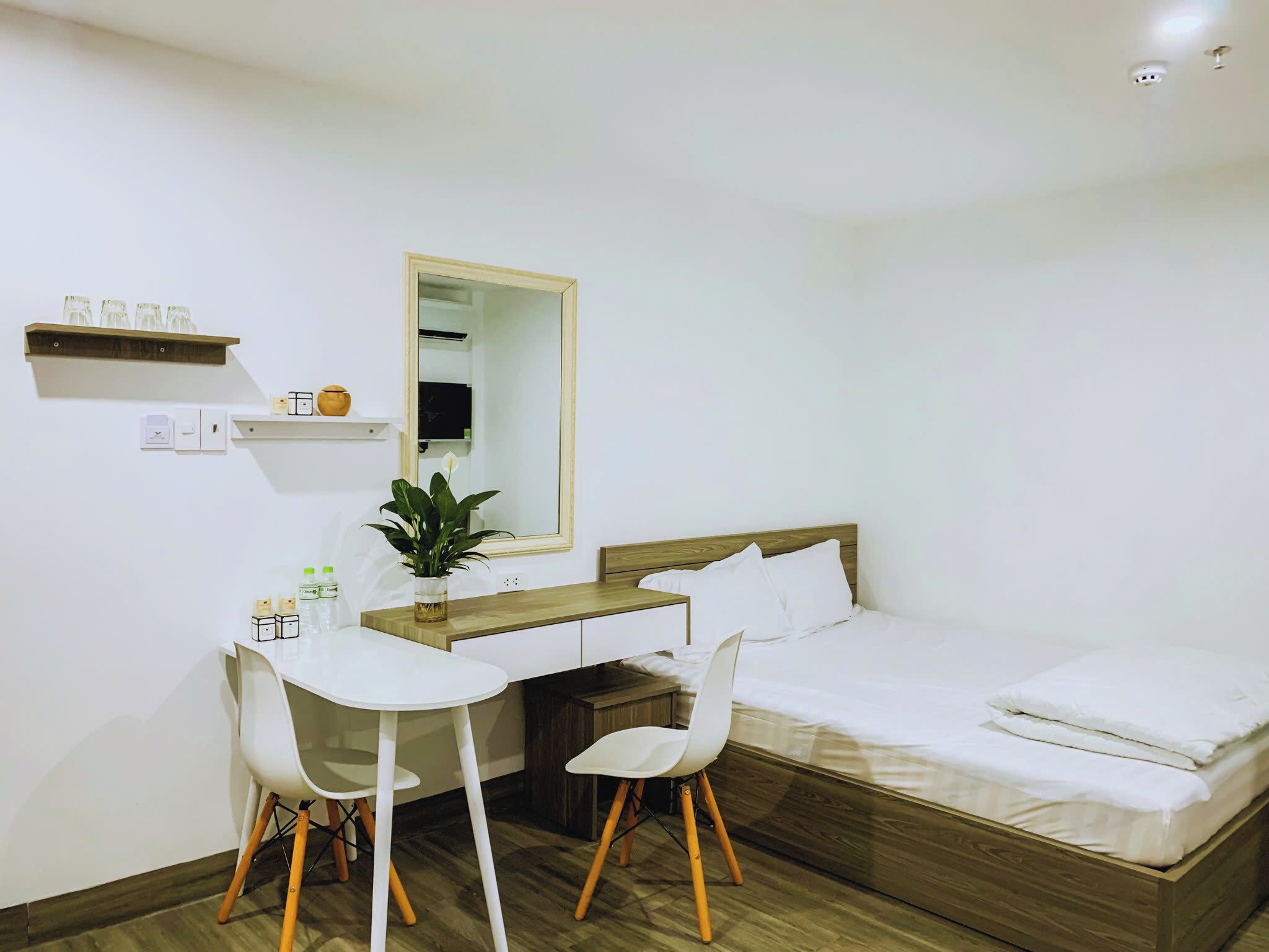 A bedroom with a wooden bed, white bedding, a desk with two white chairs, a mirror, and a small shelf with items.