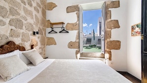 Een slaapkamer met een stenen muur, een bed met wit beddengoed, een houten hoofdeinde en uitzicht op buiten.