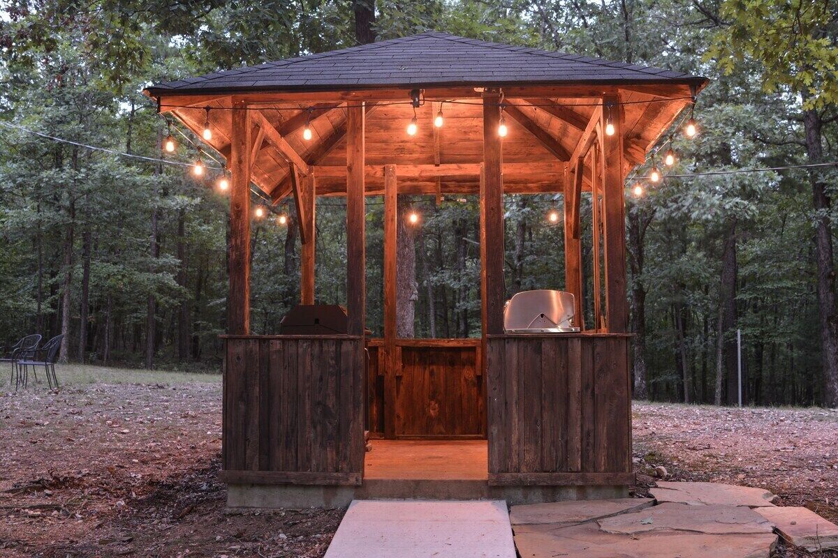 Un kiosque en bois avec un toit en métal, des guirlandes lumineuses et un barbecue.